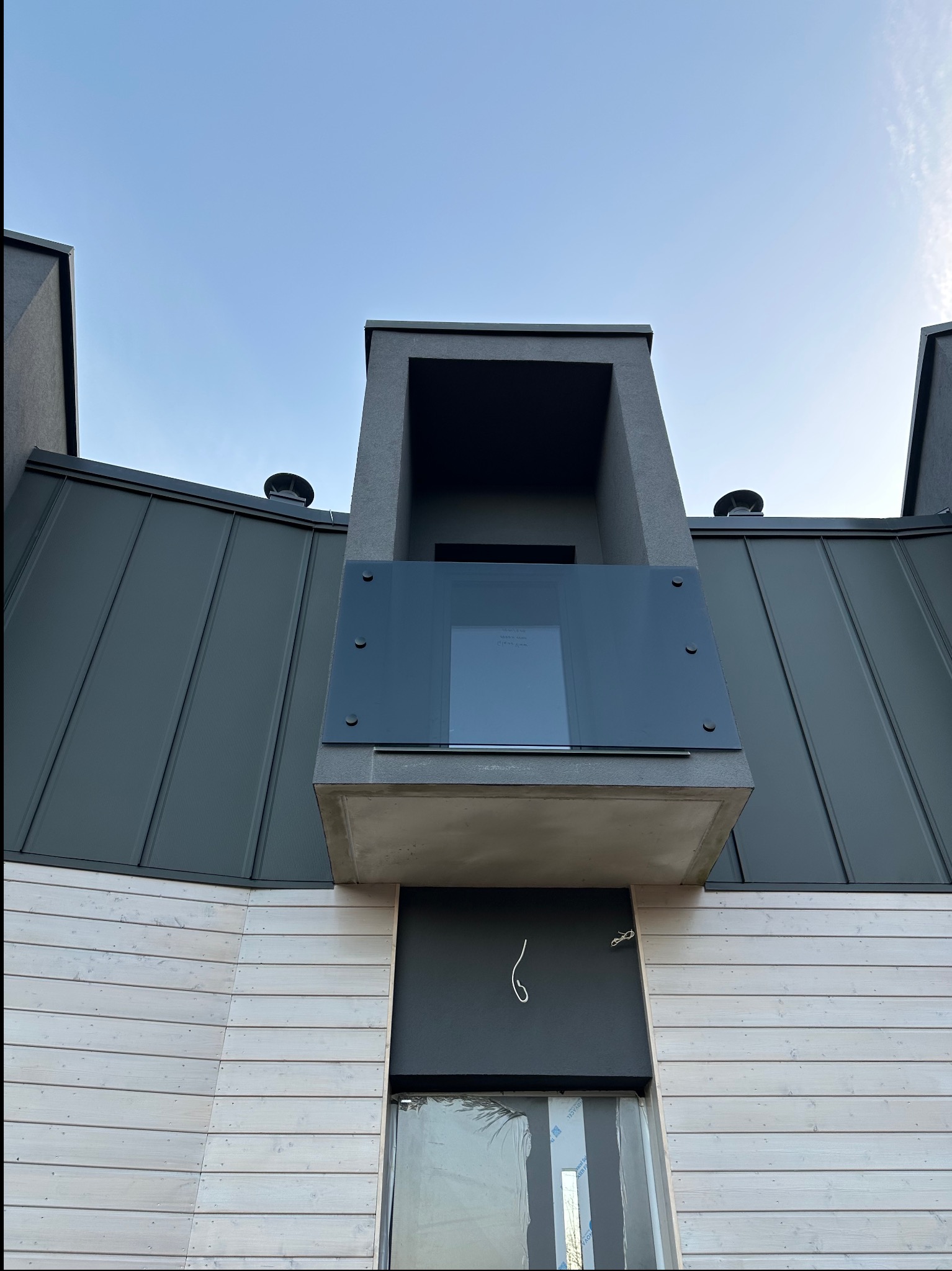 Nowoczesny balkon z szklaną balustradą, widok z dołu, na tle szarej blachy dachowej i jasnej drewnianej elewacji. Minimalistyczny design.