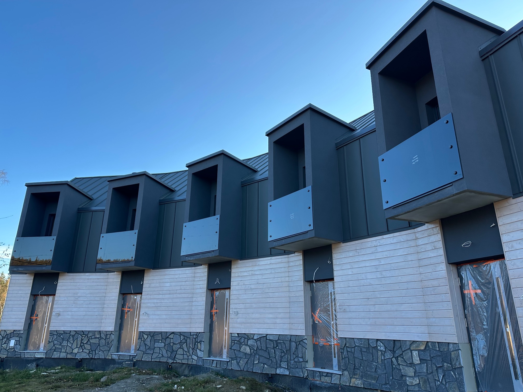 Nowoczesna elewacja budynku z balkonami z  przezroczystymi panelami. Wykończenie z kamienia i drewna. Stolarka okienna zabezpieczona folią ochronną.