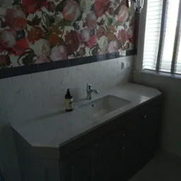 Umywalka w łazience z szarą szafką, marmurowym blatem, baterią i dozownikiem mydła na tle ściany w kwiaty i białych płytek.