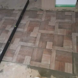 Układanie brązowych płytek podłogowych w geometryczny wzór, z użyciem krzyżyków dystansowych, widoczna poziomica i fragment surowej posadzki.