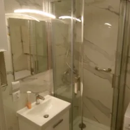 Nowoczesna kabina prysznicowa ze szklanymi drzwiami i marmurowymi płytkami na ścianach, widoczna umywalka z szafką i lustro z oświetleniem LED.