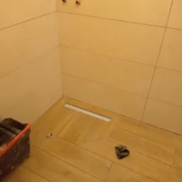 Ułożone płytki podłogowe imitujące drewno w kabinie prysznicowej z odpływem liniowym, widoczne przyłącza wodne na ścianie i wiadro z narzędziami.