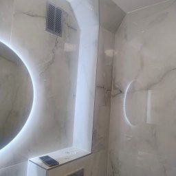 Wykończona łazienka z marmurowymi płytkami, okrągłym lustrem z podświetleniem LED i ukrytą spłuczką. Na półce leży telefon i kołki montażowe.