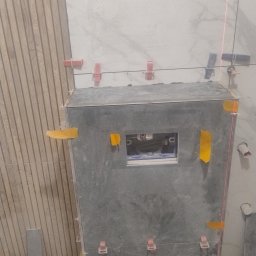 Montaż płytek w łazience: szare płytki na zabudowie stelaża WC, widoczne elementy instalacji i poziomica laserowa. Użyte klipsy poziomujące. Częściowo ułożona okładzina.