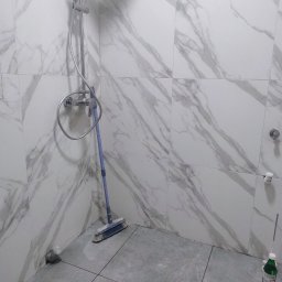 Usługi remontowo-budowlane Krzysztof Socha - Wykończona kabina prysznicowa z marmurowymi płytkami ściennymi i szarą podłogą. Widoczna bateria prysznicowa, szczotka i butelka z płynem. Minimalistyczny design.