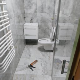 Usługi remontowo-budowlane Krzysztof Socha - Wykończona łazienka z szarymi płytkami, białą armaturą i grzejnikiem. Na podłodze leży pistolet do silikonu, co sugeruje trwające prace wykończeniowe.