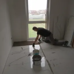 Glazurnik w trakcie układania białych płytek imitujących marmur w pomieszczeniu z oknem, widoczne narzędzia i wiaderka z klejem.