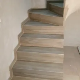 Schody wyłożone płytkami imitującymi drewno, z widocznymi niedoskonałościami na ścianach i podłodze, wskazującymi na trwający proces wykończeniowy.