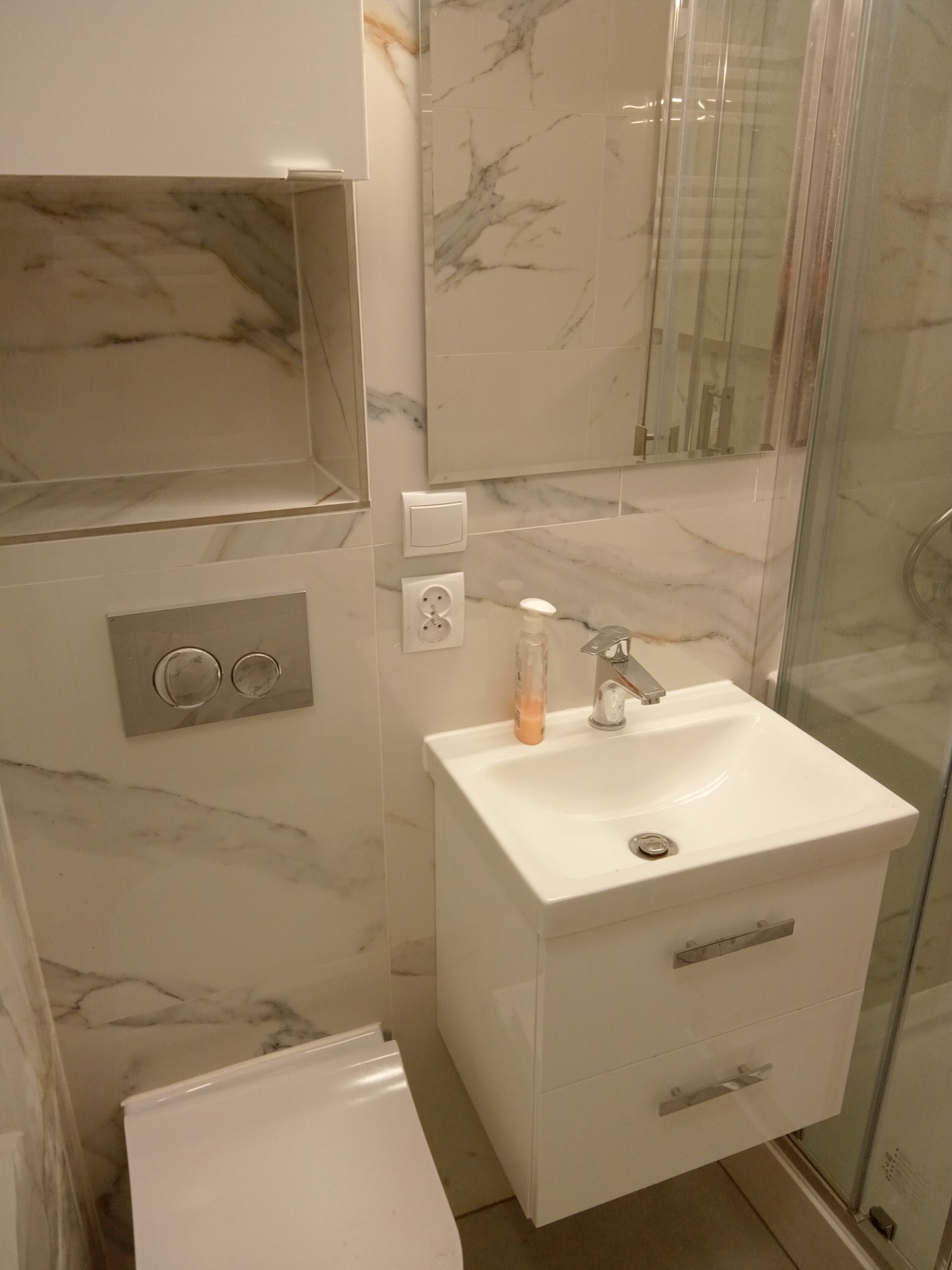 Nowoczesna łazienka z marmurowymi płytkami, białą szafką pod umywalką z dwiema szufladami, chromowaną baterią, dozownikiem mydła i wiszącą półką.
