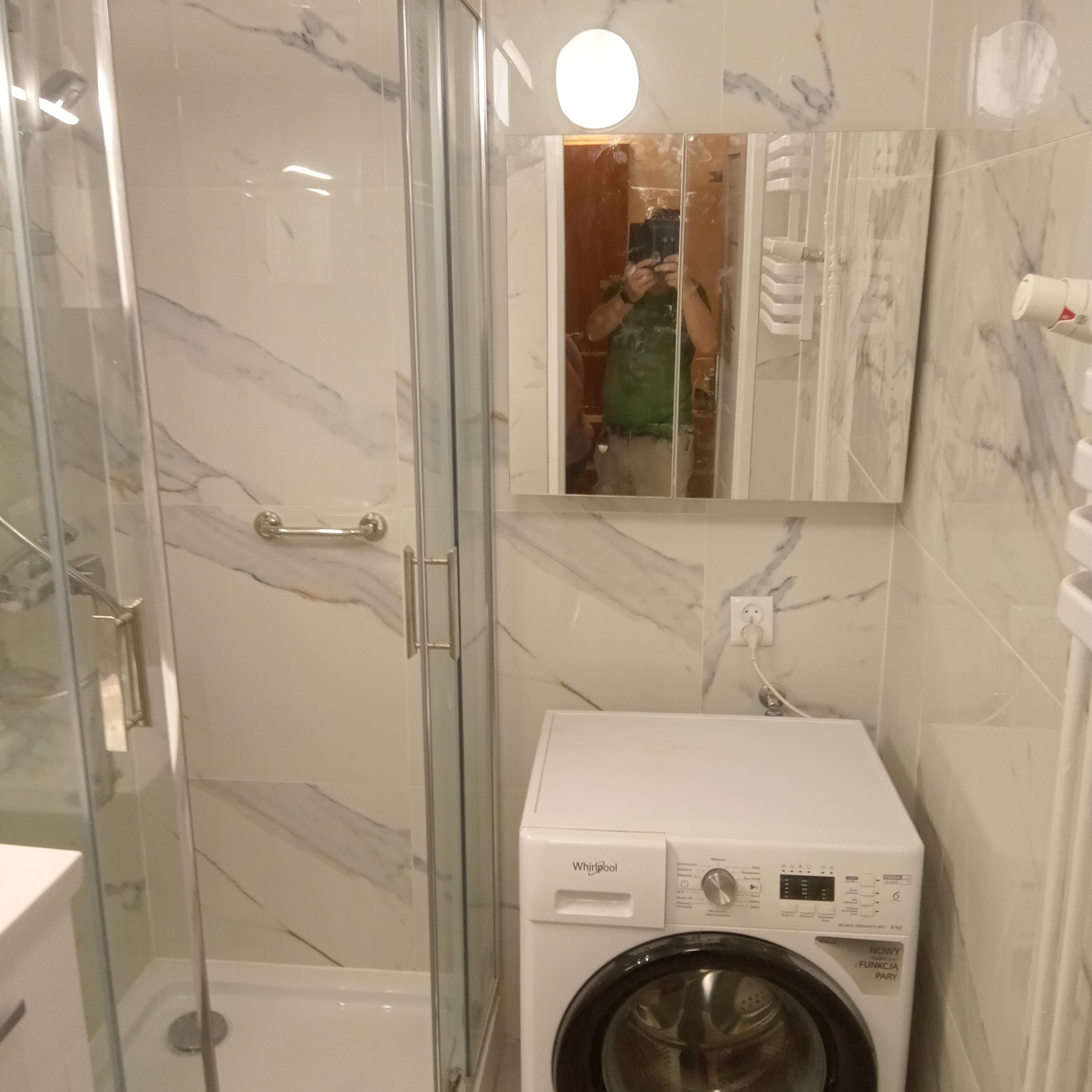 Nowoczesna łazienka z kabiną prysznicową typu walk-in i pralką Whirlpool, marmurowe płytki na ścianach, odbicie fotografa w lustrzanej szafce.