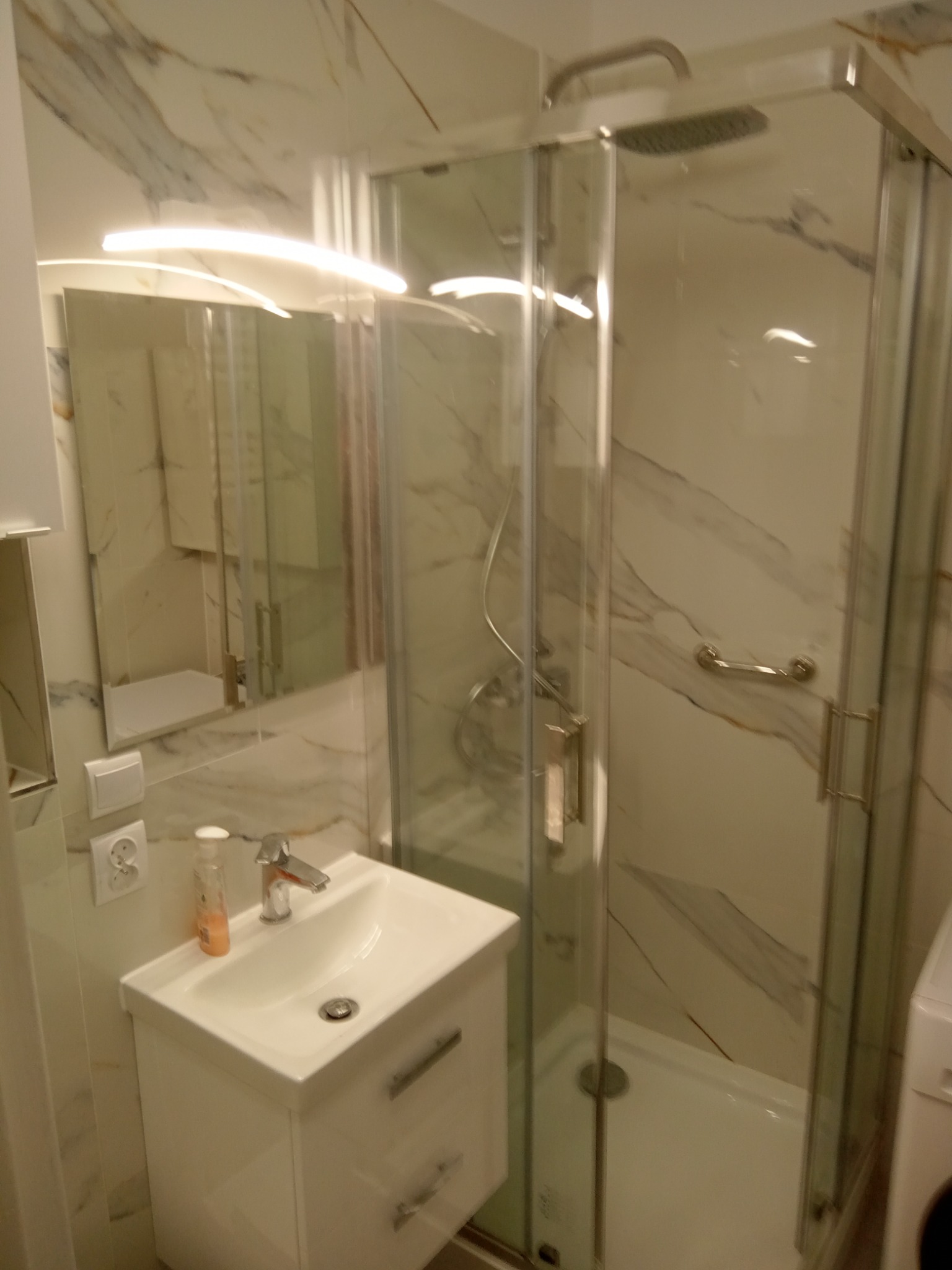 Nowoczesna kabina prysznicowa ze szklanymi drzwiami i marmurowymi płytkami na ścianach, widoczna umywalka z szafką i lustro z oświetleniem LED.