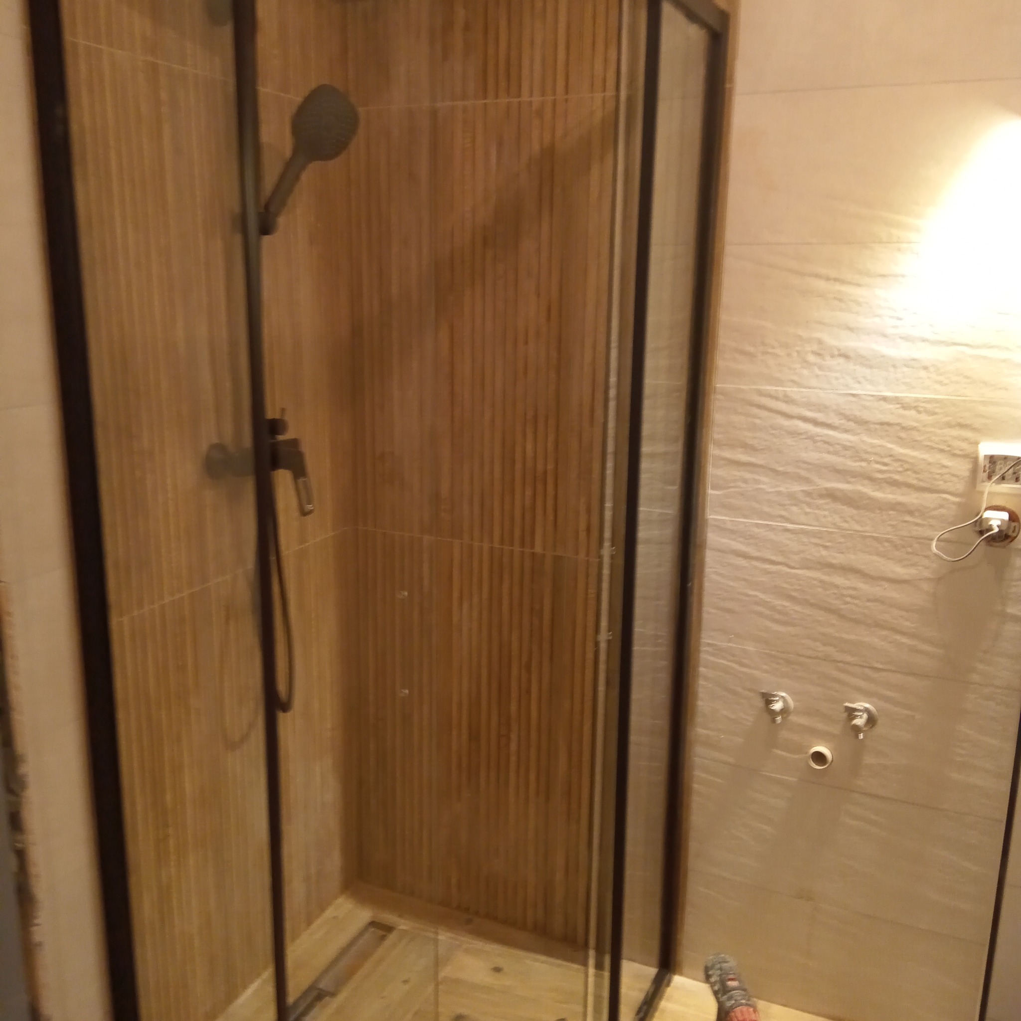 Kabina prysznicowa z czarnymi profilami i armaturą w łazience wykończonej płytkami imitującymi drewno, widoczne przyłącza hydrauliczne