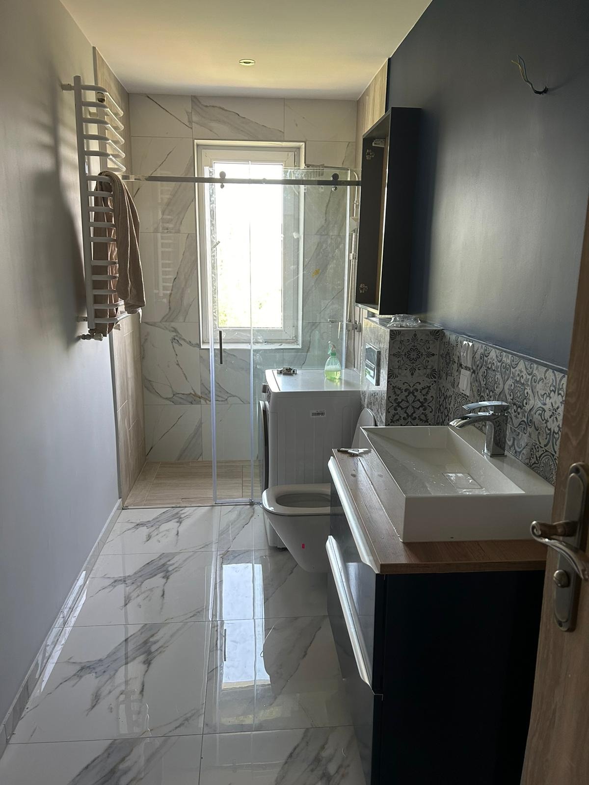 Nowoczesna łazienka z marmurowymi płytkami, kabiną prysznicową typu walk-in, grzejnikiem drabinkowym, umywalką nablatową na szafce w kolorze granatowym oraz pralką pod oknem.