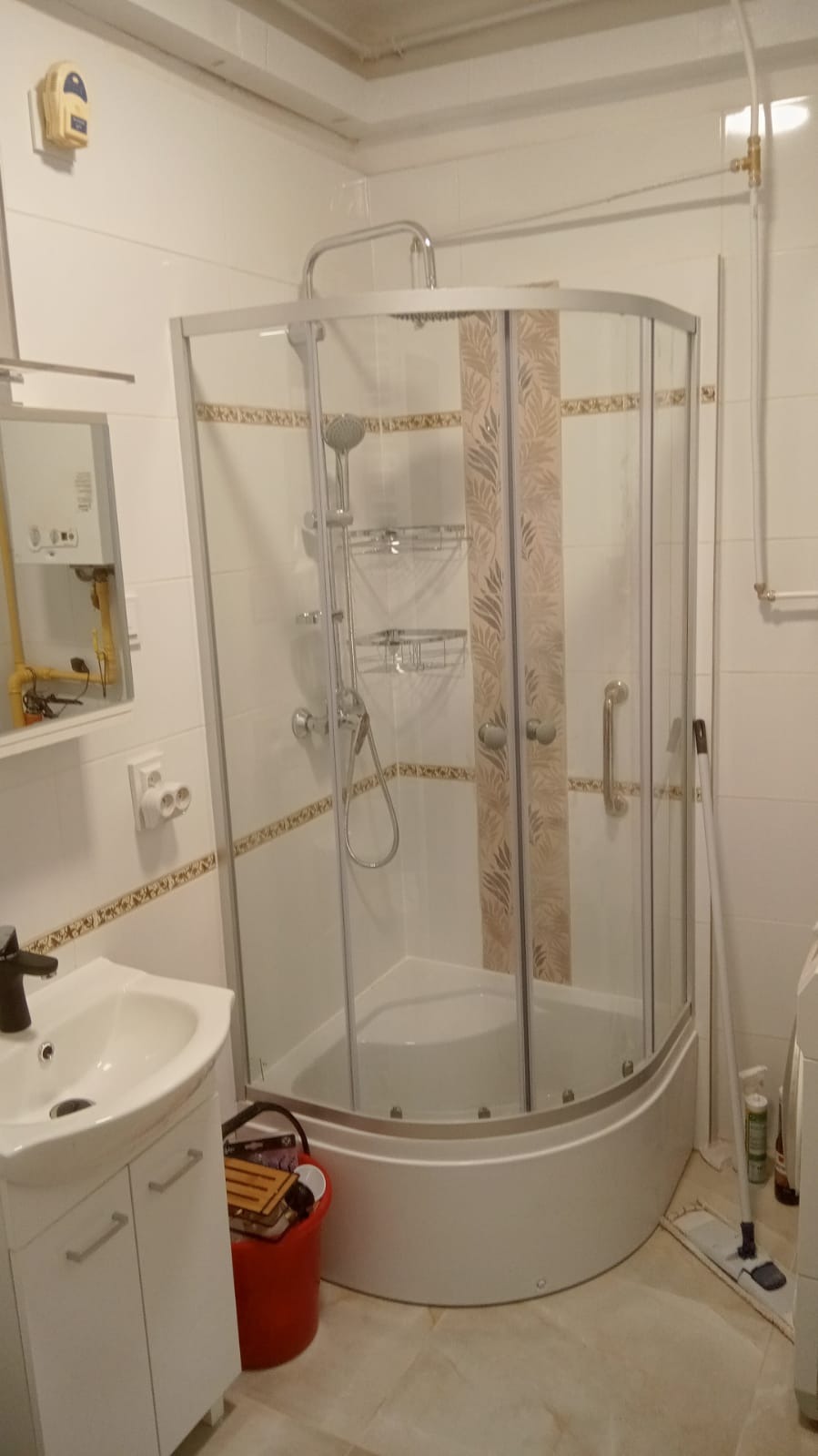 Narożna kabina prysznicowa z wzorem liści na panelach, umywalka z szafką i widoczny bojler gazowy w łazience z beżowymi płytkami podłogowymi.