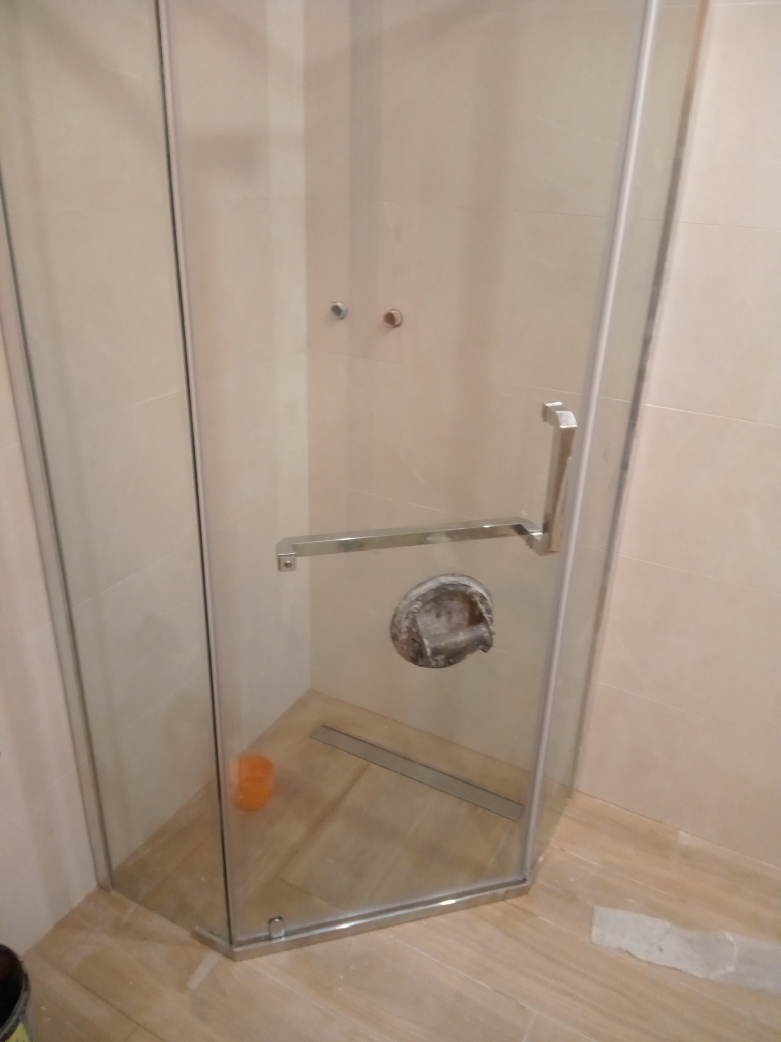 Narożna kabina prysznicowa z chromowanymi okuciami, widoczny odpływ liniowy i niedoczyszczona armatura, jasne płytki imitujące drewno.