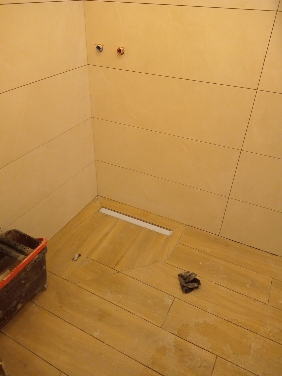 Ułożone płytki podłogowe imitujące drewno w kabinie prysznicowej z odpływem liniowym, widoczne przyłącza wodne na ścianie i wiadro z narzędziami.