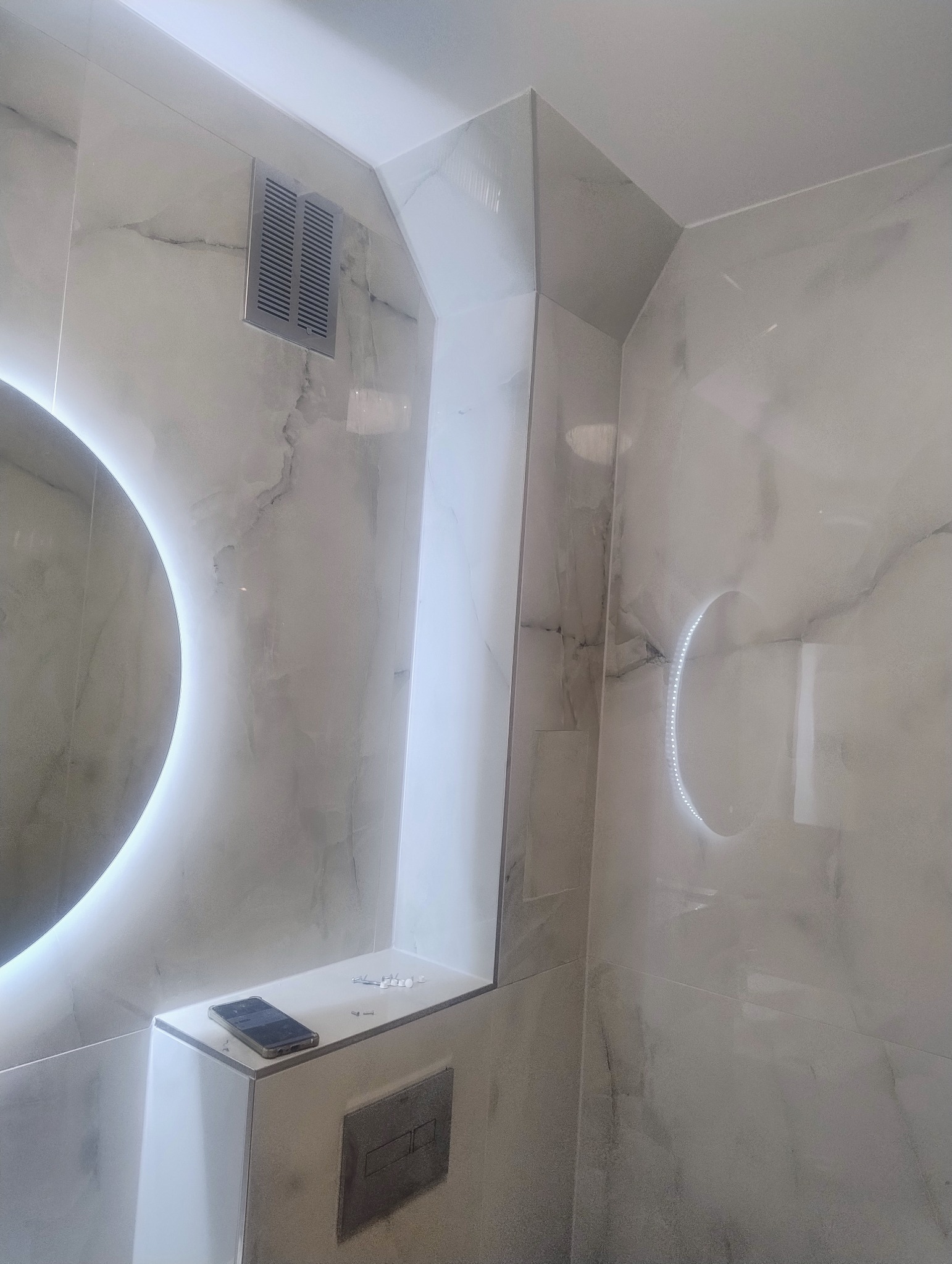 Wykończona łazienka z marmurowymi płytkami, okrągłym lustrem z podświetleniem LED i ukrytą spłuczką. Na półce leży telefon i kołki montażowe.