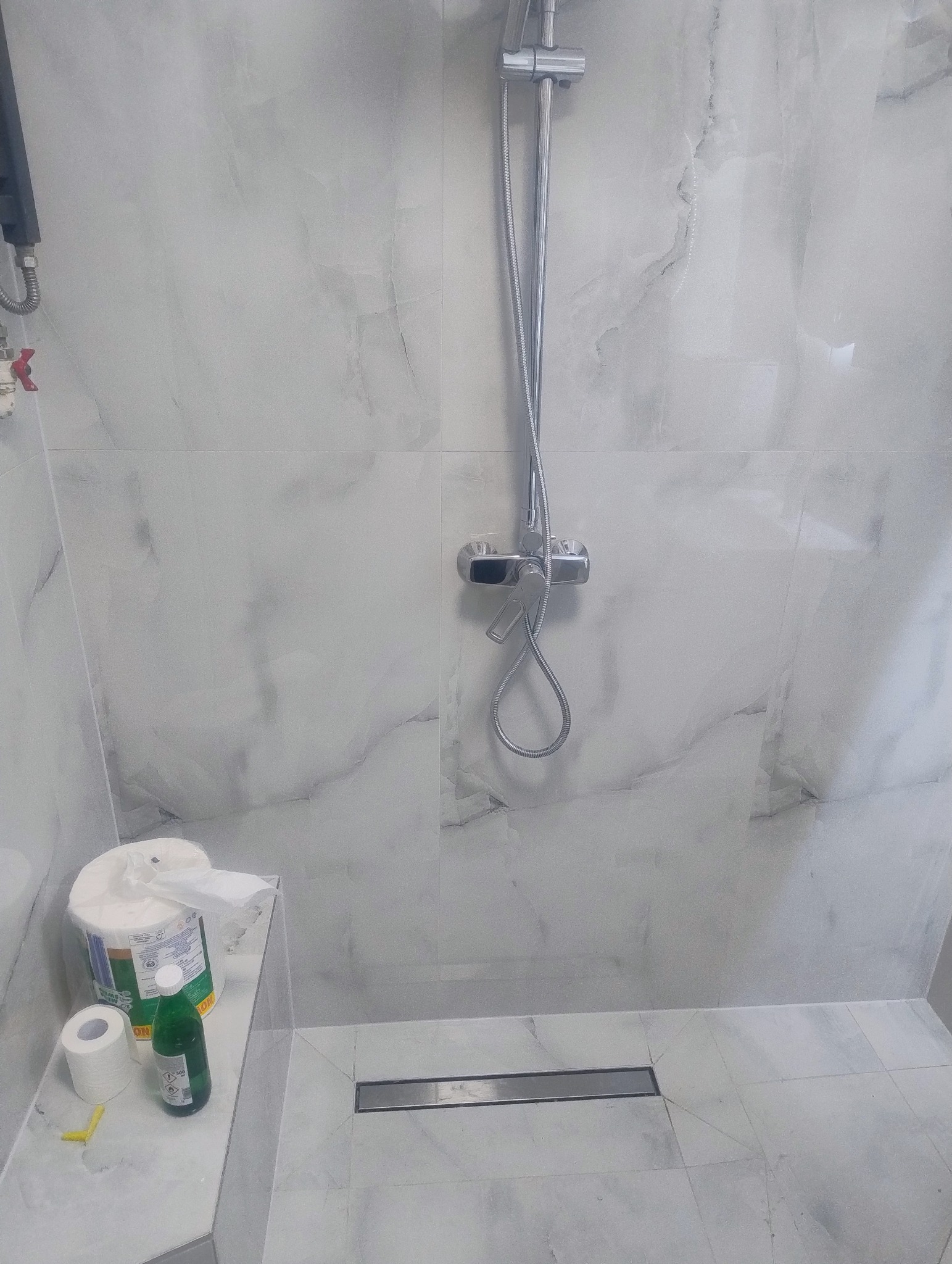 Wykończona kabina prysznicowa z odpływem liniowym i armaturą w chromie, wyłożona płytkami imitującymi marmur. Widoczne akcesoria łazienkowe.