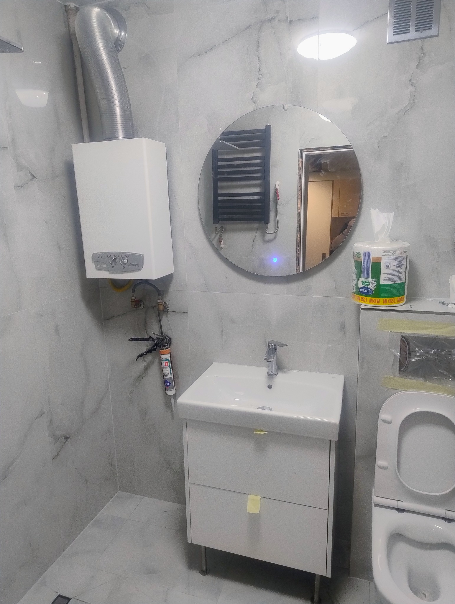 Wykończona łazienka w szarościach: biała umywalka z szafką, toaleta, okrągłe lustro z grzejnikiem. Na ścianie piec gazowy z rurą. Widoczne narzędzia i akcesoria.