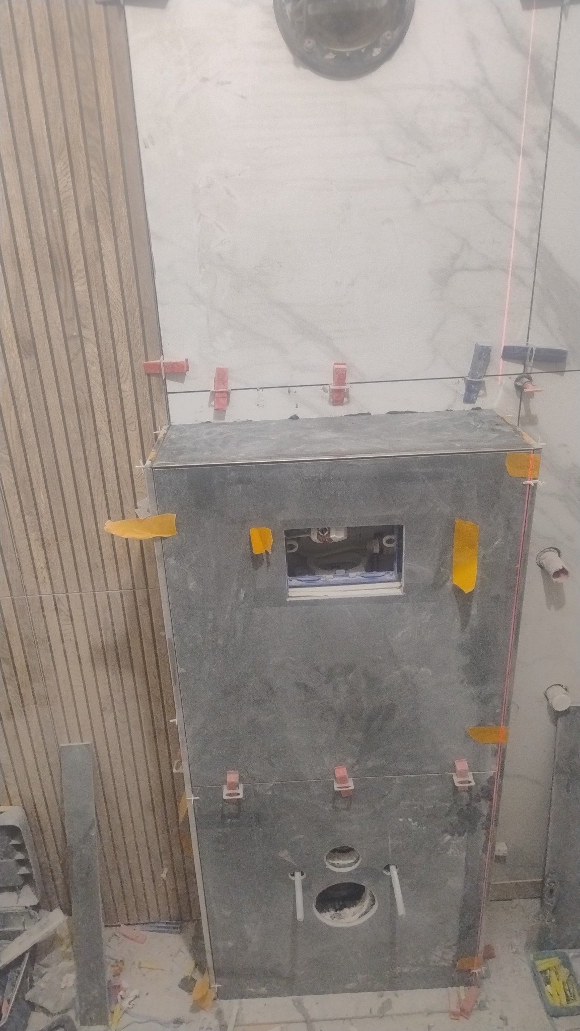 Montaż płytek w łazience: szare płytki na zabudowie stelaża WC, widoczne elementy instalacji i poziomica laserowa. Użyte klipsy poziomujące. Częściowo ułożona okładzina.