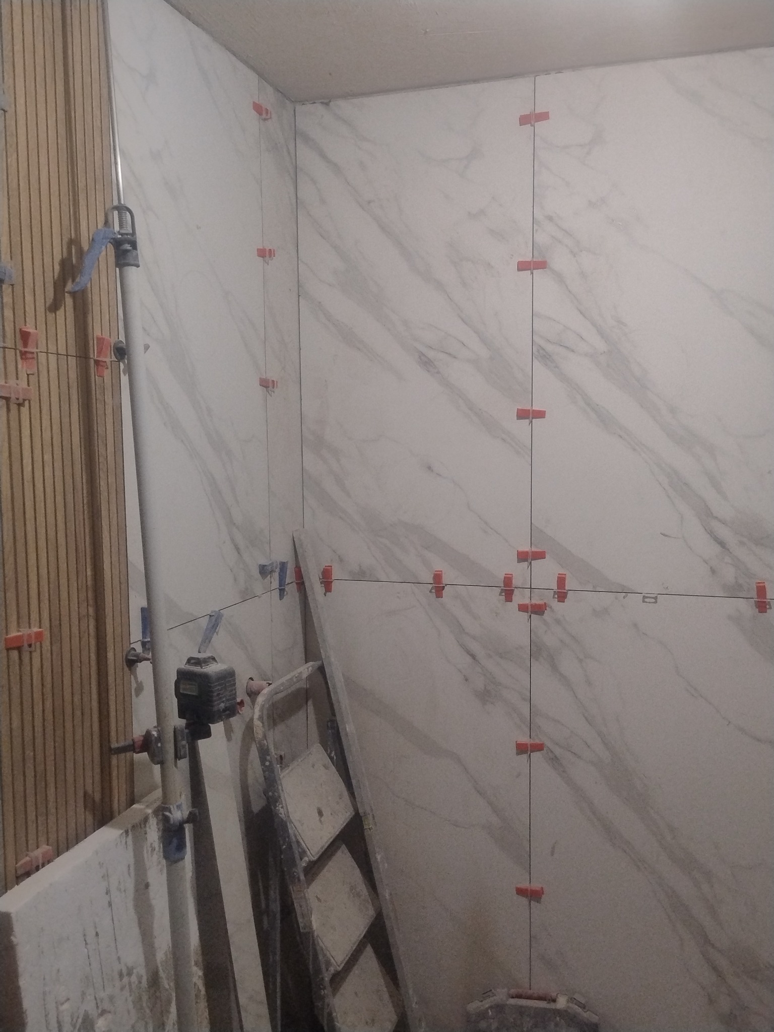 Ściana w trakcie układania płytek imitujących marmur, z użyciem poziomicy laserowej i klipsów poziomujących. Widoczny wózek glazurniczy i fragmenty drewnianej konstrukcji.