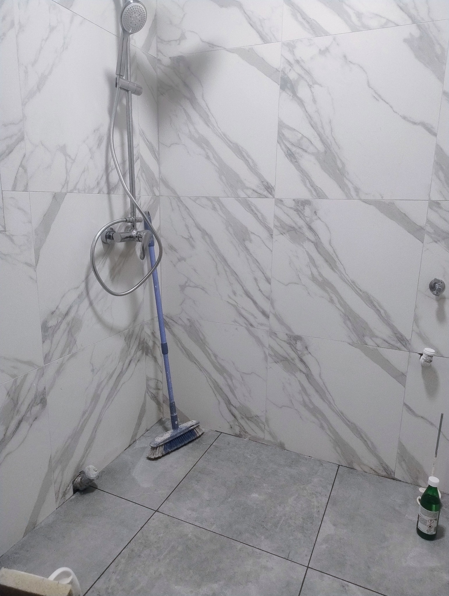 Wykończona kabina prysznicowa z marmurowymi płytkami ściennymi i szarą podłogą. Widoczna bateria prysznicowa, szczotka i butelka z płynem. Minimalistyczny design.