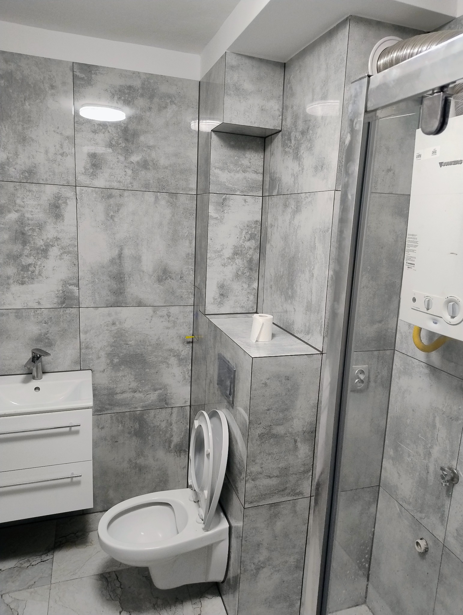 Wykończona łazienka w szarościach z kabiną prysznicową, umywalką z szafką i toaletą z otwartą klapą. Na ścianach duże, kwadratowe płytki imitujące beton.