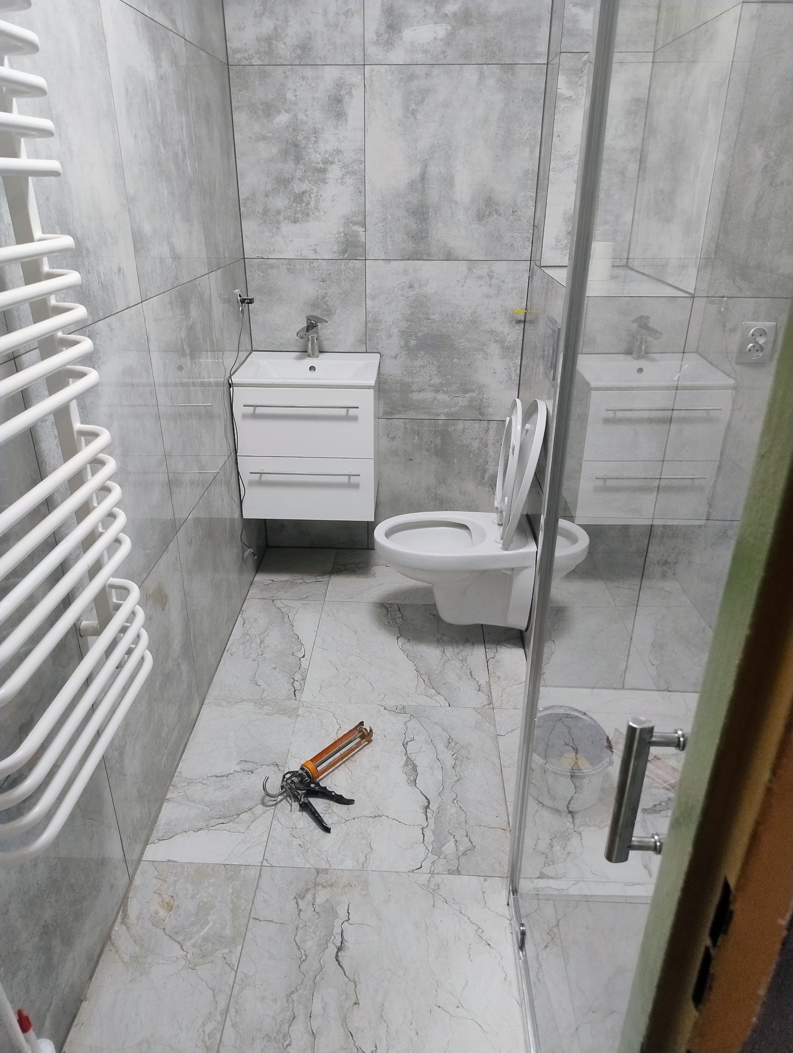 Wykończona łazienka z szarymi płytkami, białą armaturą i grzejnikiem. Na podłodze leży pistolet do silikonu, co sugeruje trwające prace wykończeniowe.