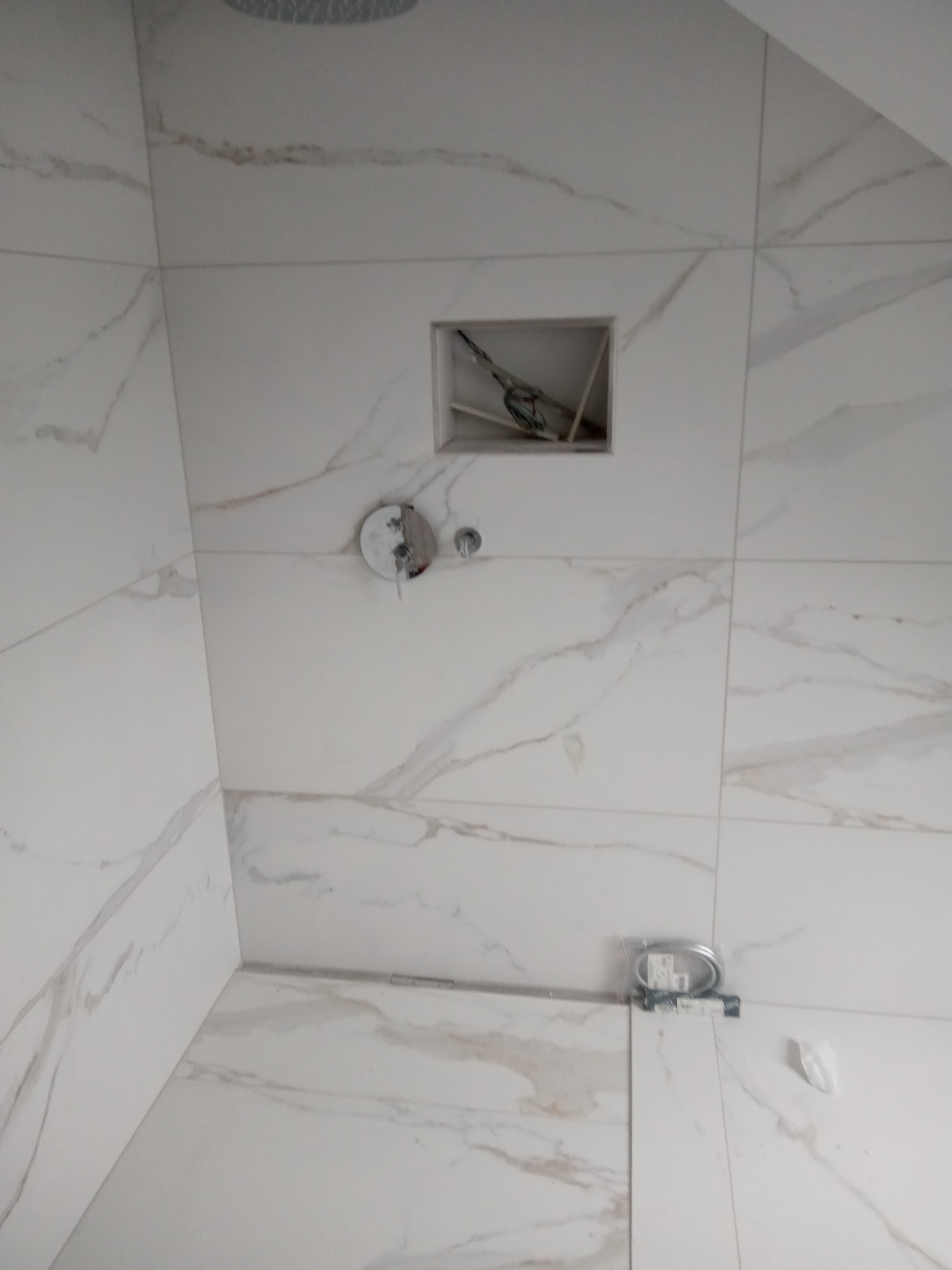 Ściana kabiny prysznicowej wyłożona białymi płytkami imitującymi marmur, z widoczną wnęką w ścianie z wystającymi przewodami, zamontowaną baterią prysznicową i pakietem węży prysznicowych na podłodze.