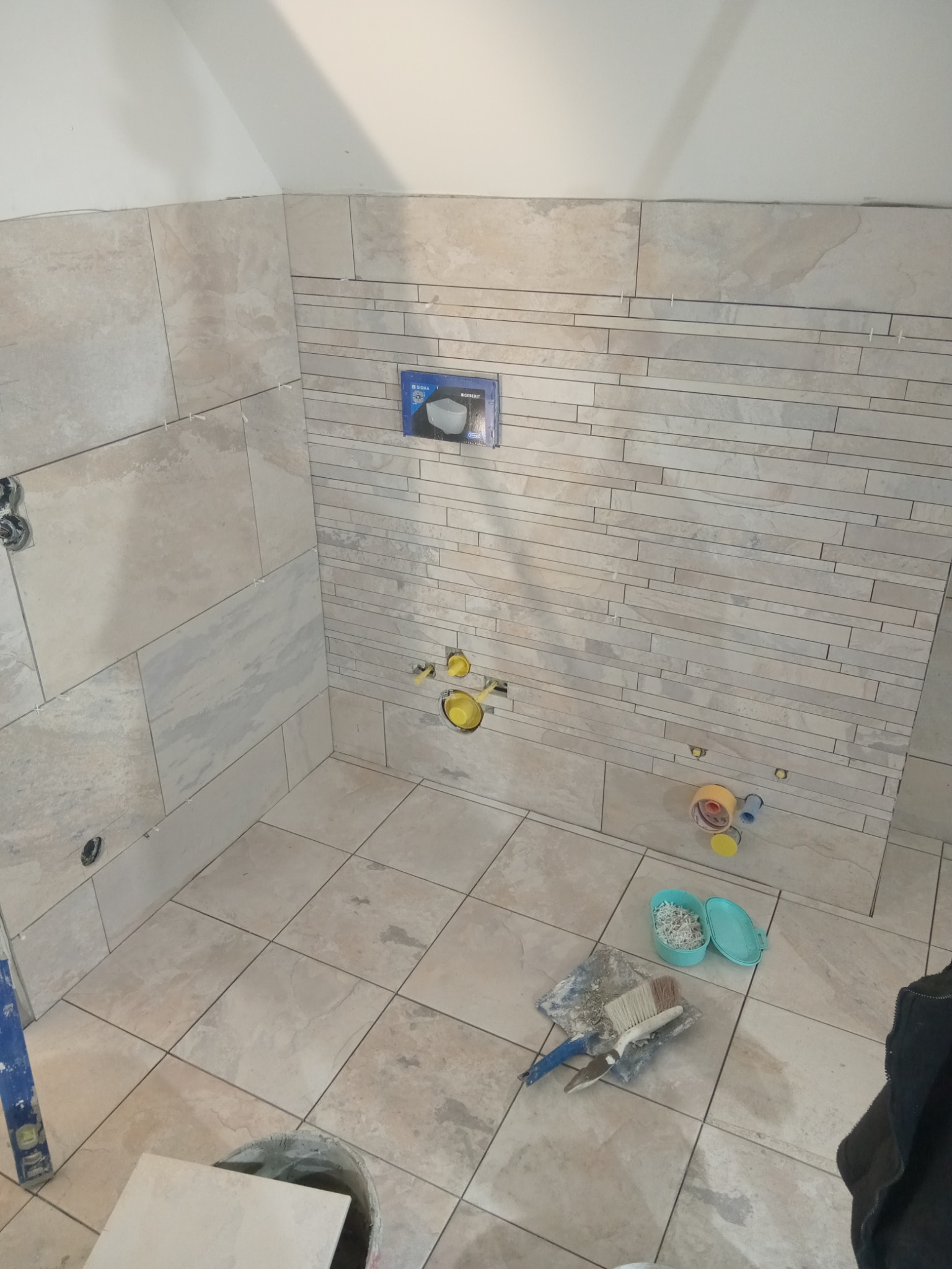 Ściana łazienki częściowo wyłożona beżowymi płytkami w dwóch różnych wzorach, widoczne narzędzia glazurnicze i instalacja podtynkowa WC