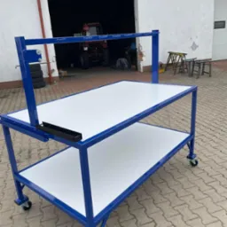 Wielopoziomowy, mobilny stół warsztatowy z niebieskiej stali i białymi blatami, na tle otwartego garażu z widocznym traktorem.