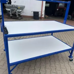 Stół warsztatowy na kółkach, rama metalowa pomalowana na niebiesko, dwa blaty w kolorze białym, tło: budynek gospodarczy, samochód dostawczy i maszyny rolnicze.