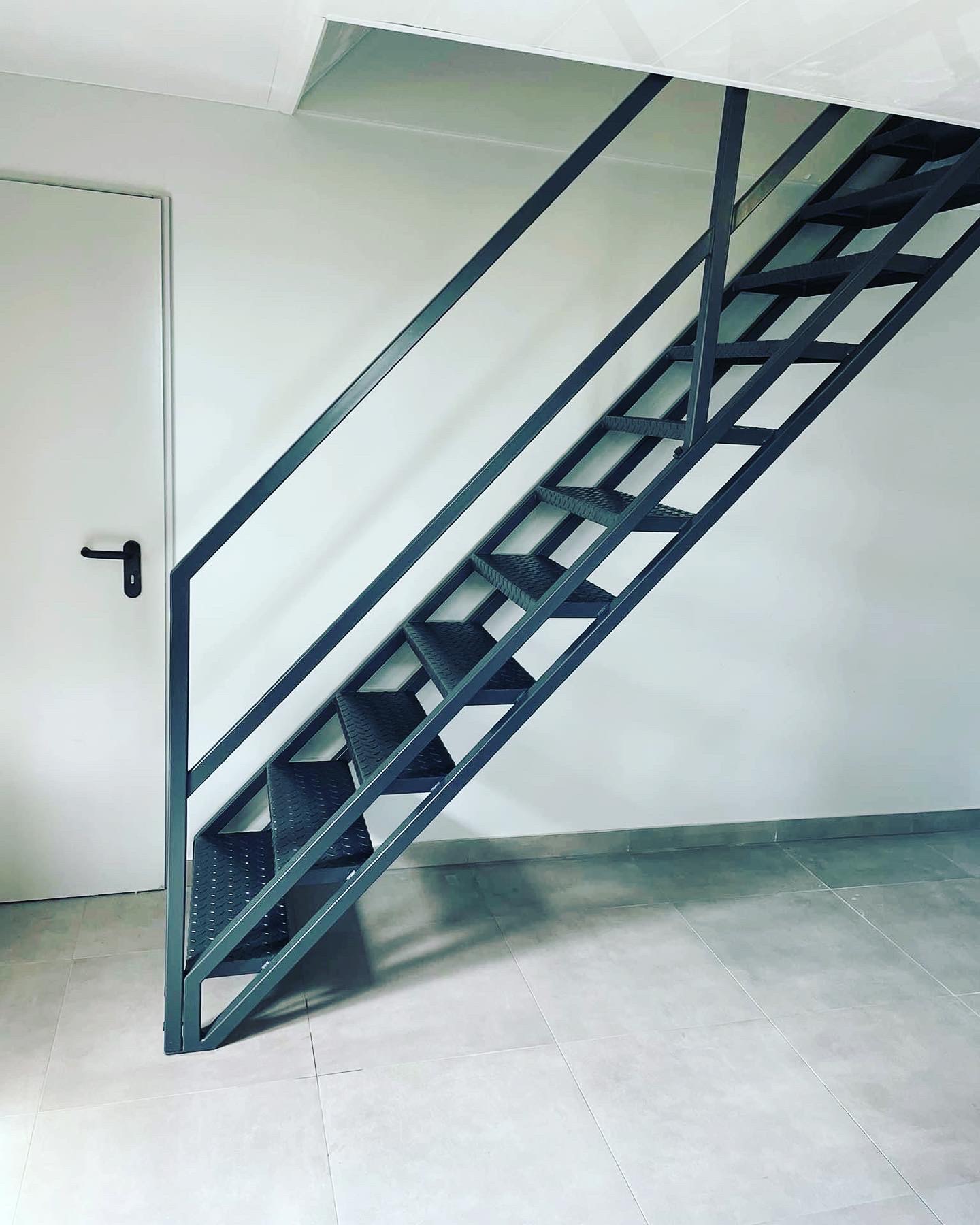 Szare metalowe schody ażurowe z perforowanymi stopniami, widok z dołu na tle białej ściany i drzwi