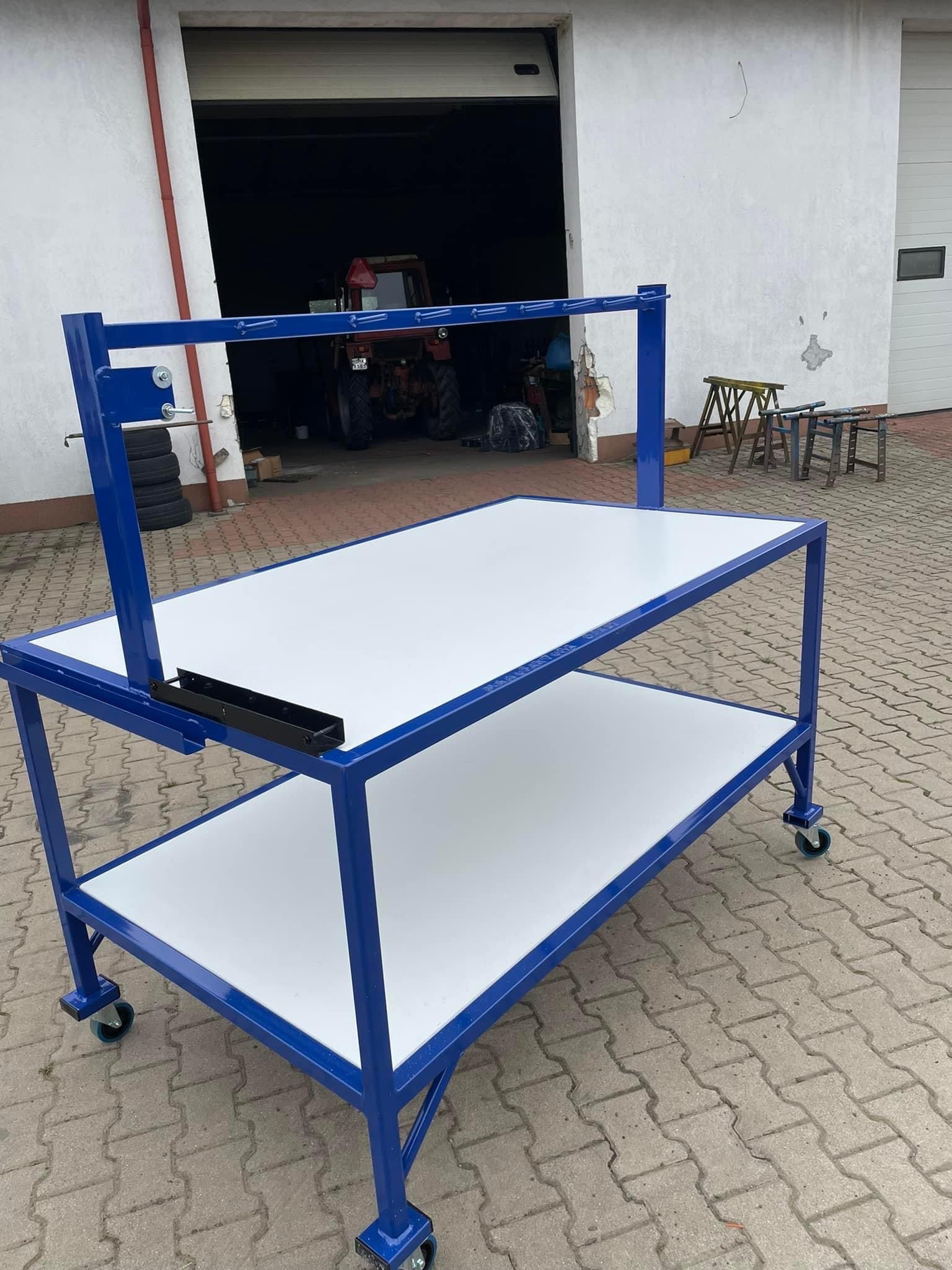 Wielopoziomowy, mobilny stół warsztatowy z niebieskiej stali i białymi blatami, na tle otwartego garażu z widocznym traktorem.