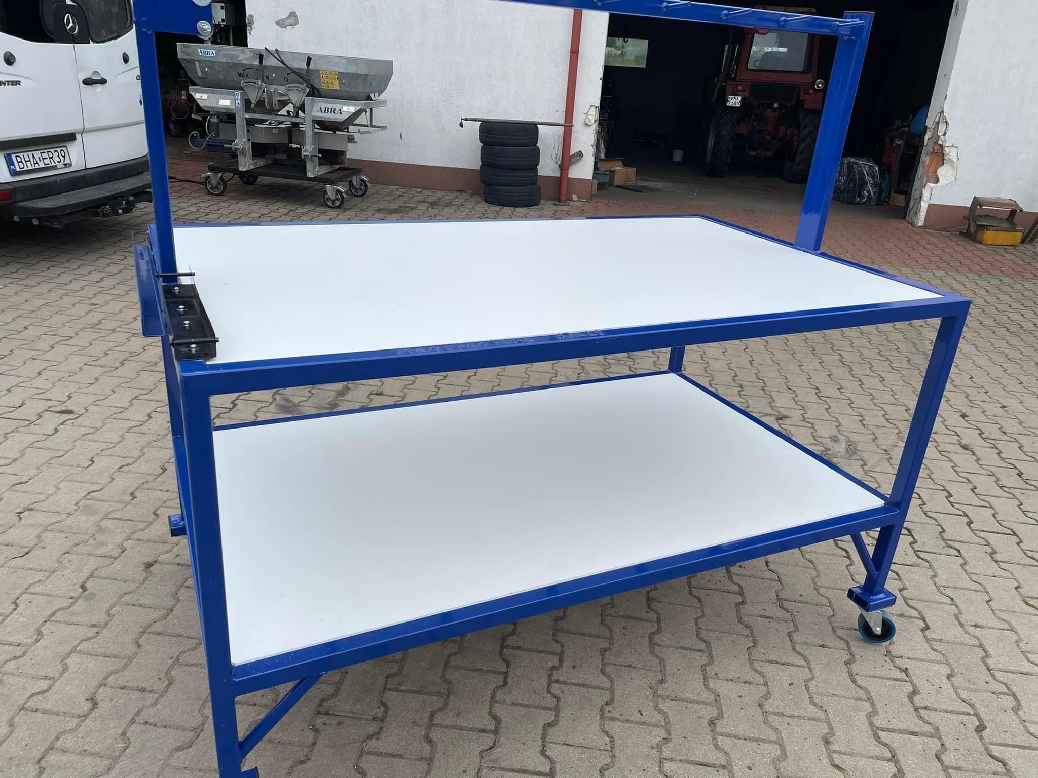 Stół warsztatowy na kółkach, rama metalowa pomalowana na niebiesko, dwa blaty w kolorze białym, tło: budynek gospodarczy, samochód dostawczy i maszyny rolnicze.