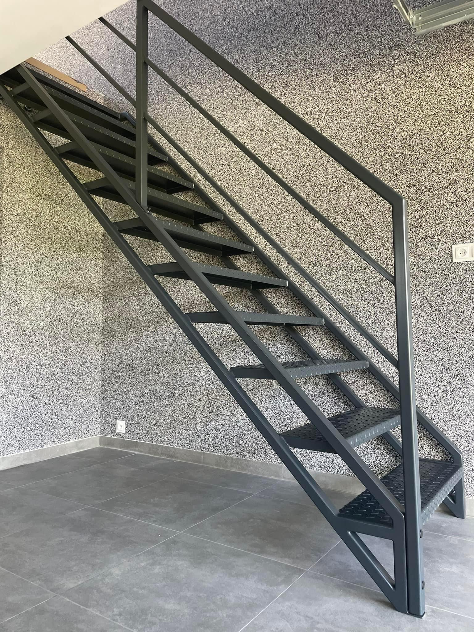 Szare metalowe schody wewnętrzne z poręczą, widok z dołu, na tle ściany z fakturą i szarej podłogi z płytek.