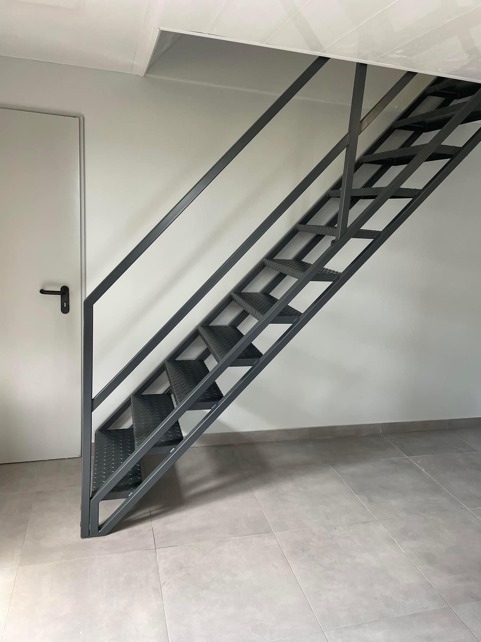Szare metalowe schody z perforowanymi stopniami, prowadzące do otworu w suficie, widoczne na tle białej ściany i drzwi z czarną klamką.