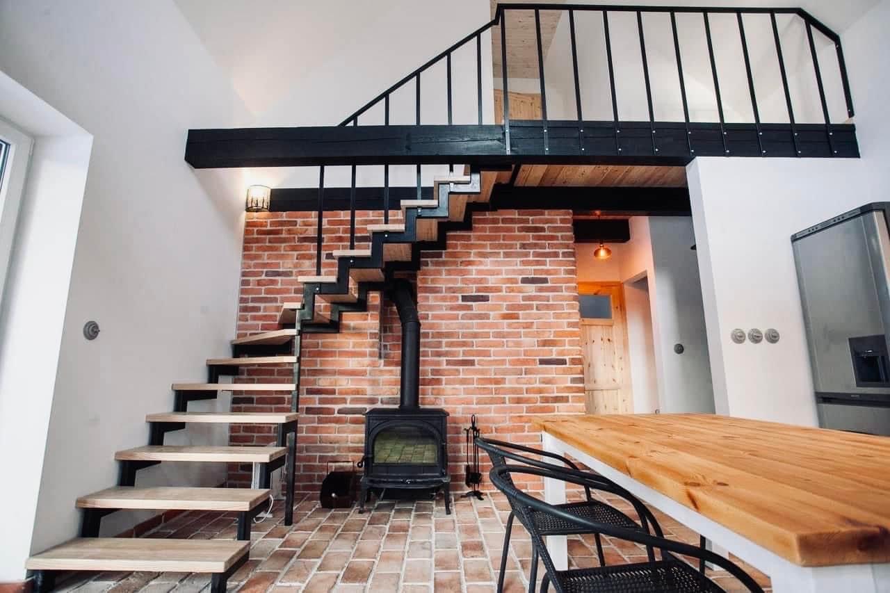 Nowoczesne schody metalowe z drewnianymi stopniami, prowadzące na antresolę z czarną balustradą, na tle ściany z czerwonej cegły i pieca wolnostojącego.