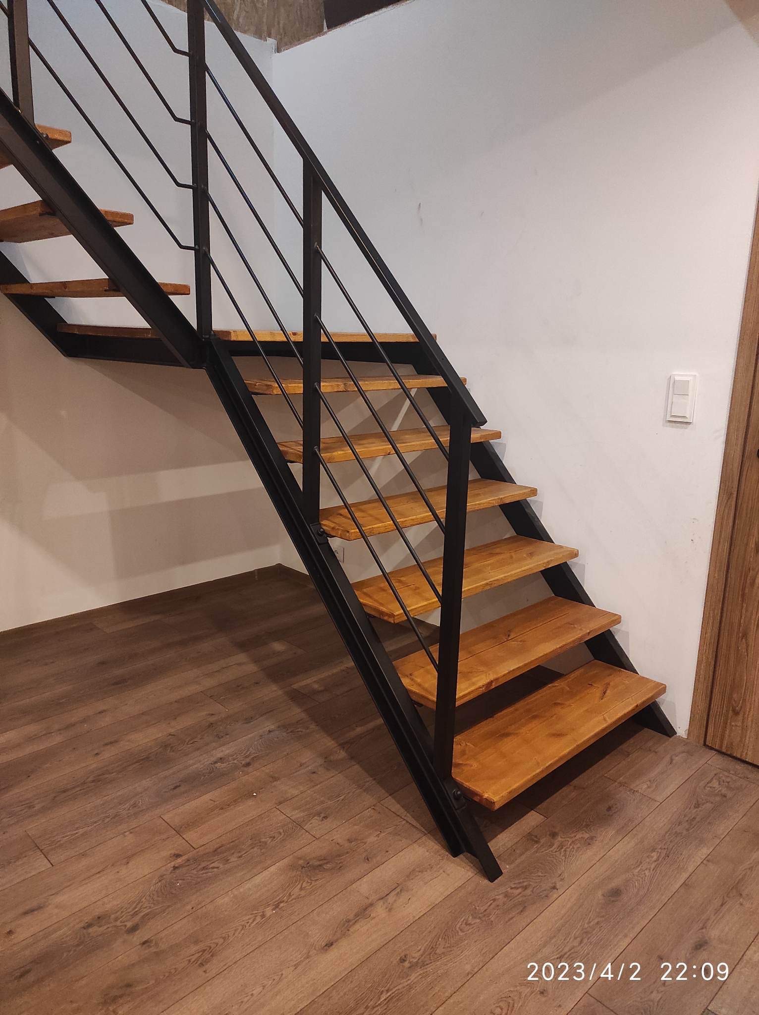 Metalowe schody z drewnianymi stopniami, czarna konstrukcja, minimalistyczny design, widok z dołu na tle drewnianej podłogi i białej ściany z włącznikiem.