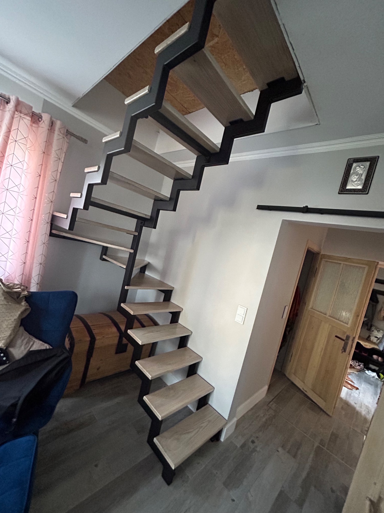 Schody metalowe z drewnianymi stopniami prowadzące na poddasze, widoczne z dołu, w tle zasłony i kufer.