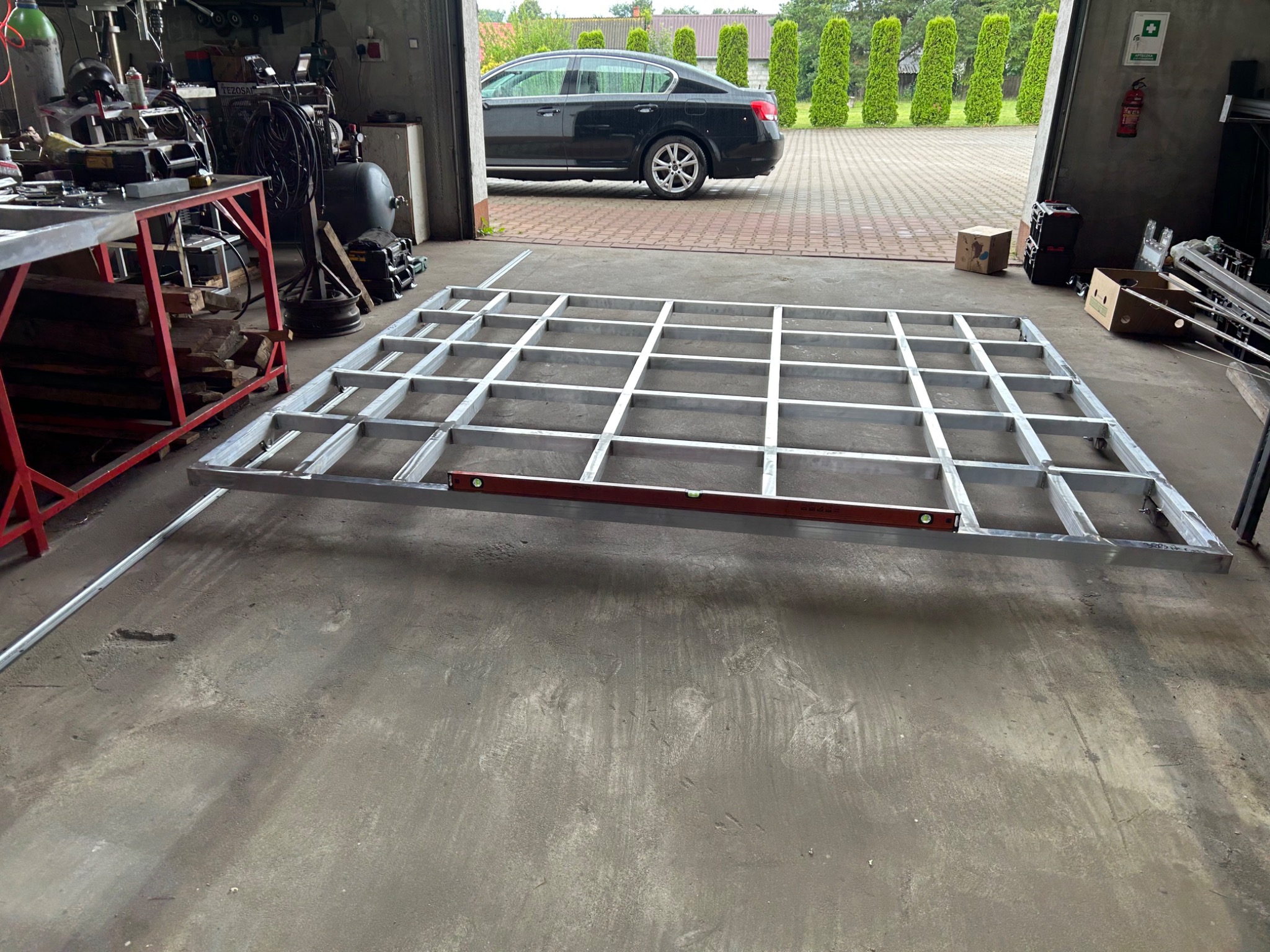 Szkielet metalowej konstrukcji kratownicowej z poziomicą, w warsztacie z widocznym samochodem i narzędziami w tle.