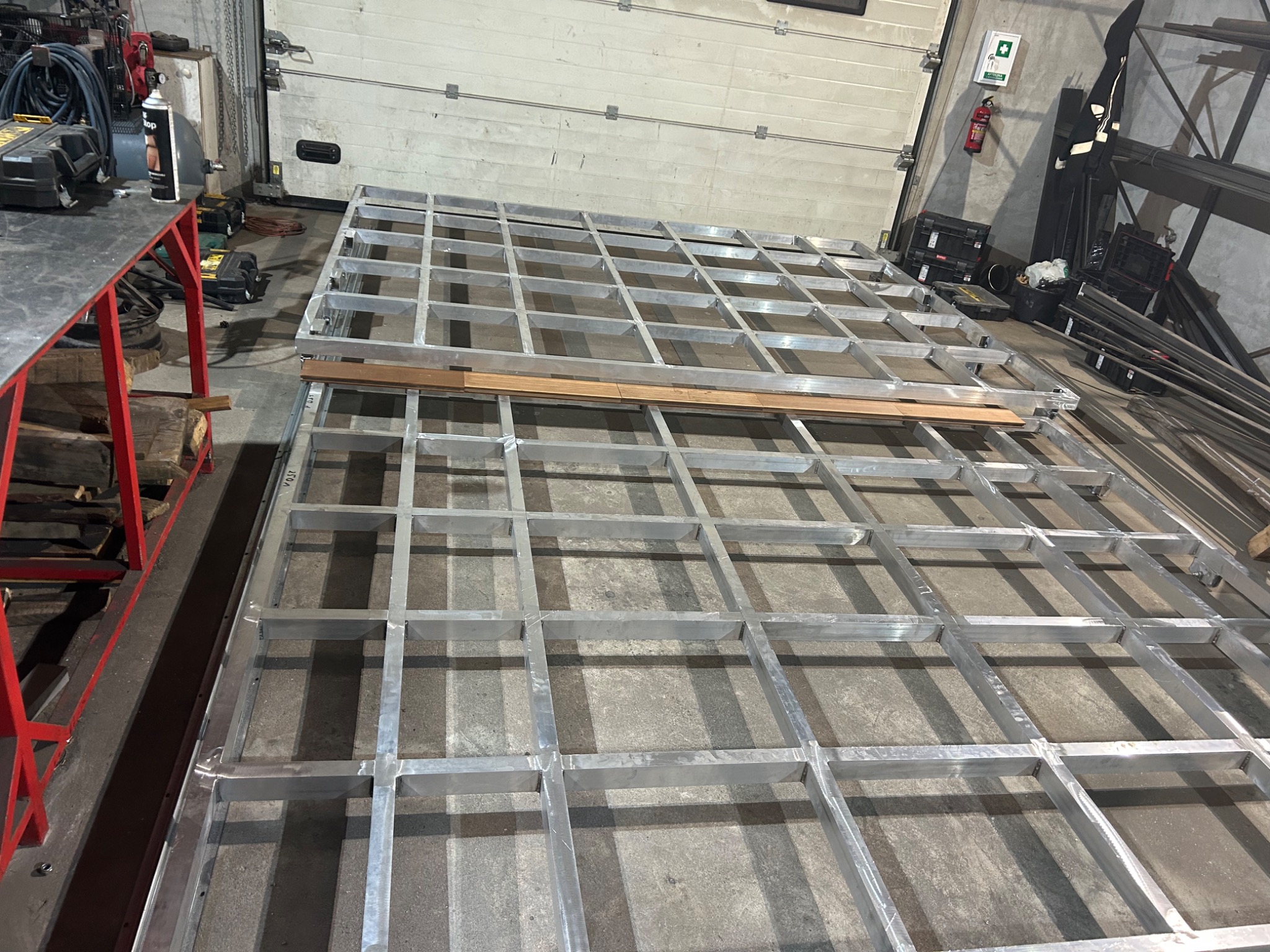 Szkielet konstrukcji metalowej z profili aluminiowych, ułożony na podłodze warsztatu, z widocznymi spawami i fragmentami drewnianych elementów.