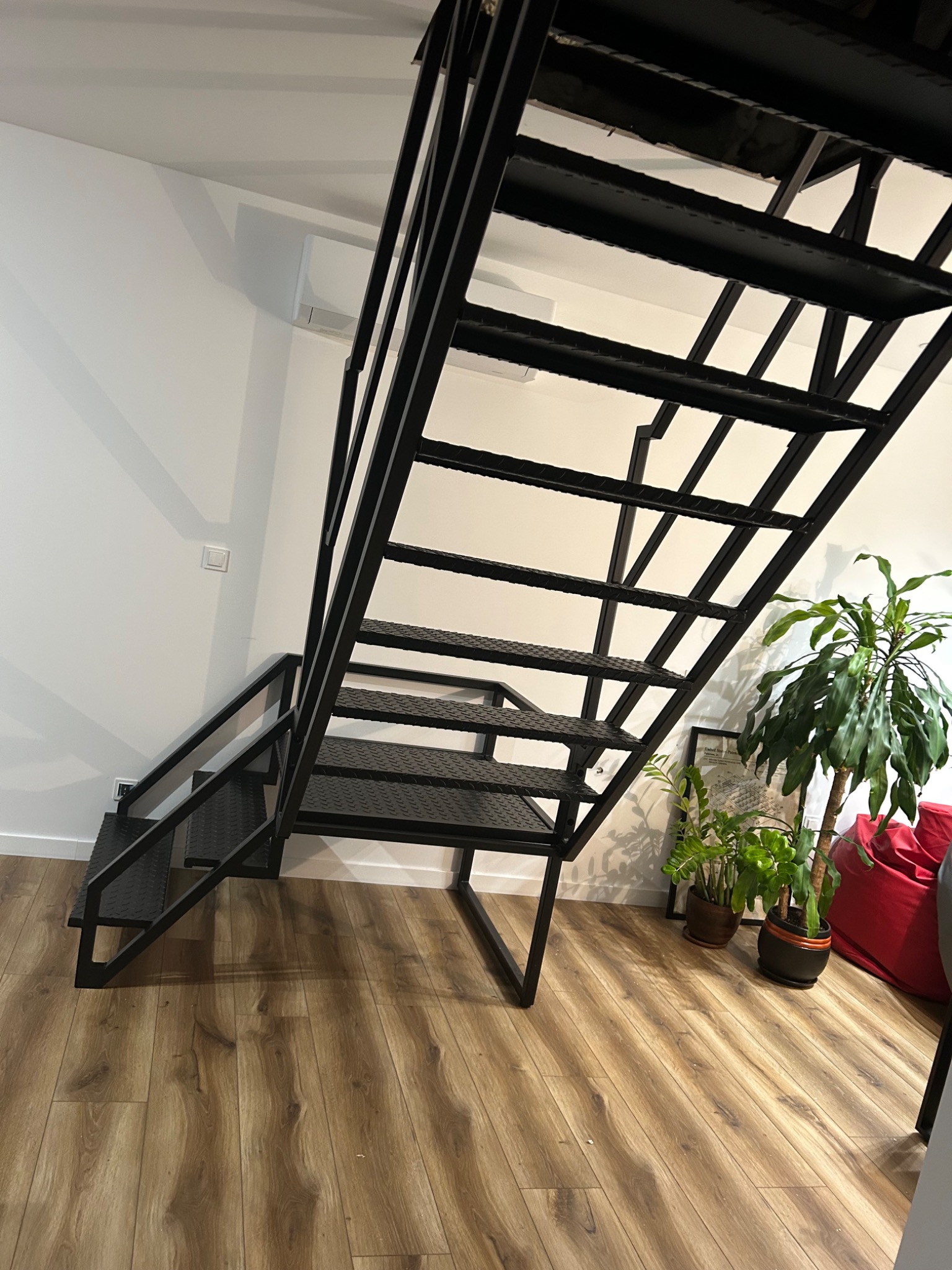 Czarne, metalowe schody o ażurowej konstrukcji w nowoczesnym wnętrzu, z widocznymi roślinami doniczkowymi i podłogą z paneli.