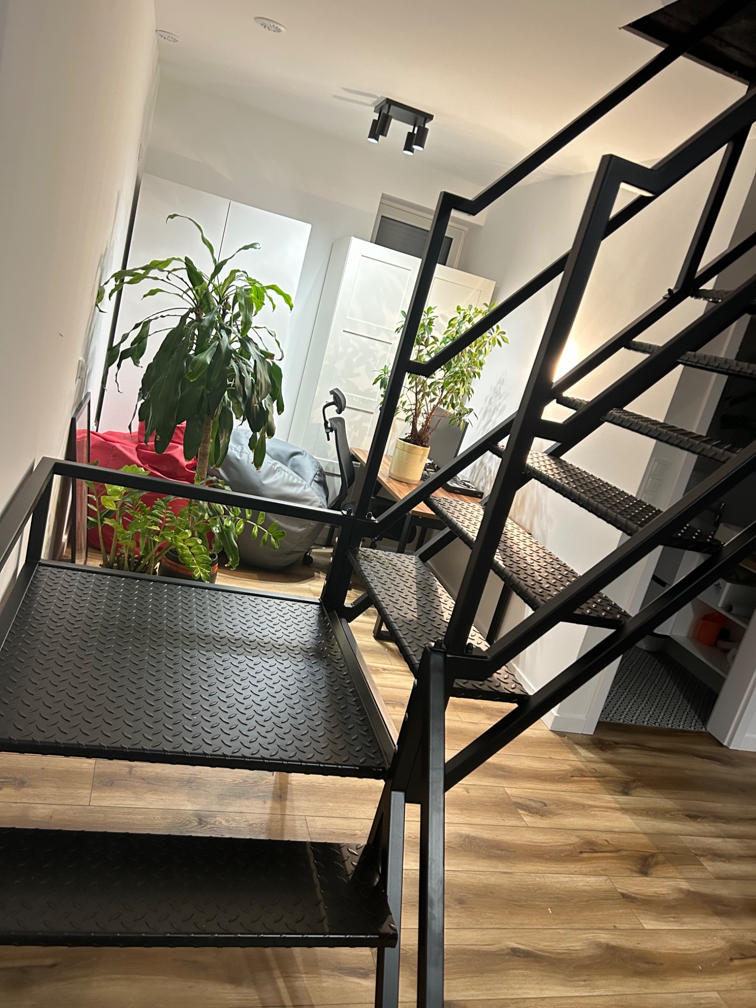 Nowoczesne schody metalowe o konstrukcji ażurowej, z czarnymi stopniami z blachy ryflowanej, w jasnym wnętrzu z drewnianą podłogą i roślinami doniczkowymi.