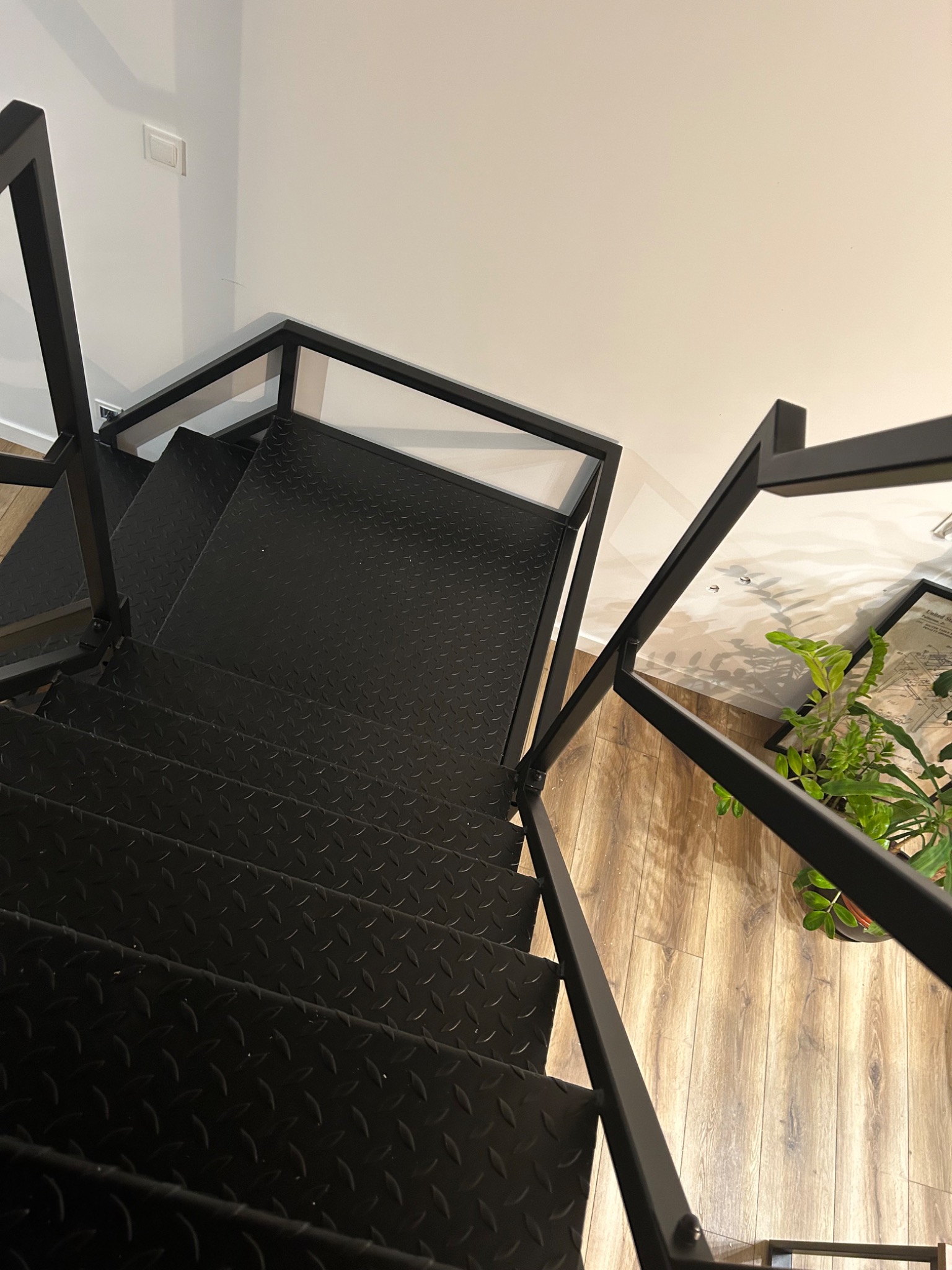 Czarno-matowe schody metalowe z perforowanymi stopniami, widok z góry, z rośliną doniczkową w tle na drewnianej podłodze.