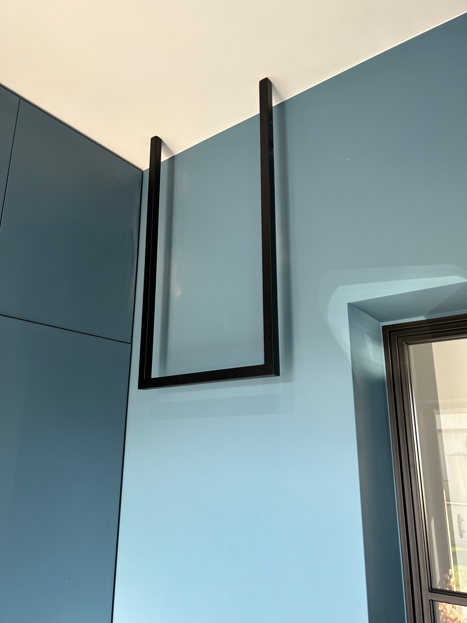 Minimalistyczna, czarna, metalowa rama podwieszona do sufitu w nowoczesnym wnętrzu z błękitnymi ścianami i szafkami.