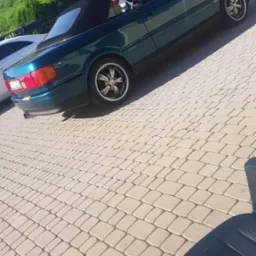 Zielony kabriolet Audi na kostce brukowej, widoczne felgi aluminiowe i inne auta w tle.