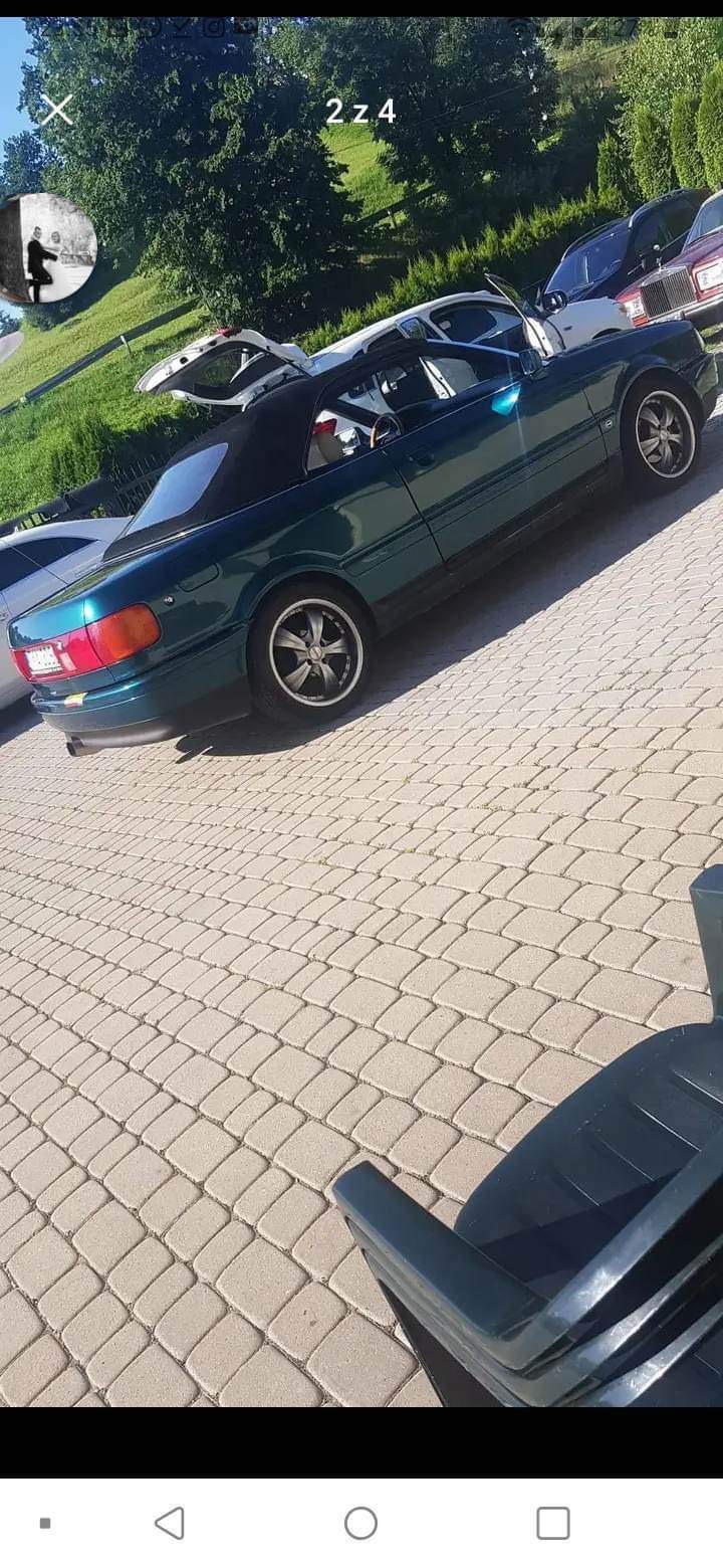 Zielony kabriolet Audi na kostce brukowej, widoczne felgi aluminiowe i inne auta w tle.