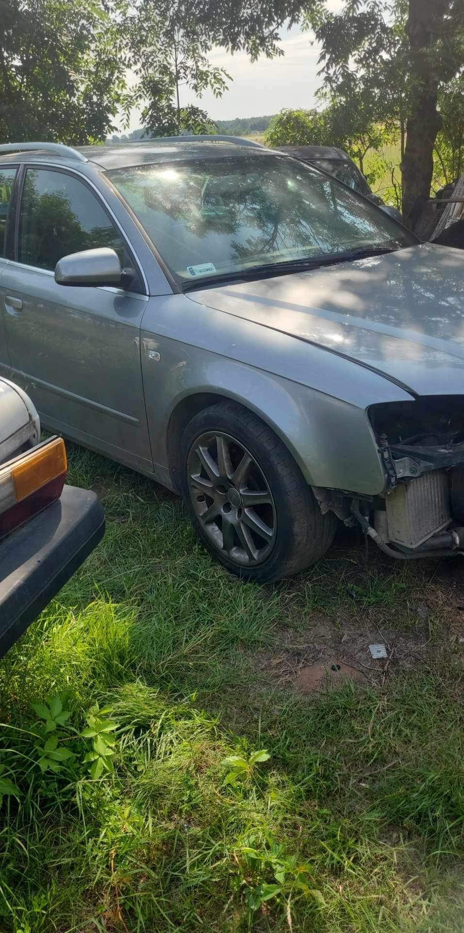 Srebrny samochód osobowy z uszkodzonym przodem, stojący na trawie, z widoczną chłodnicą, obok inny, starszy pojazd.