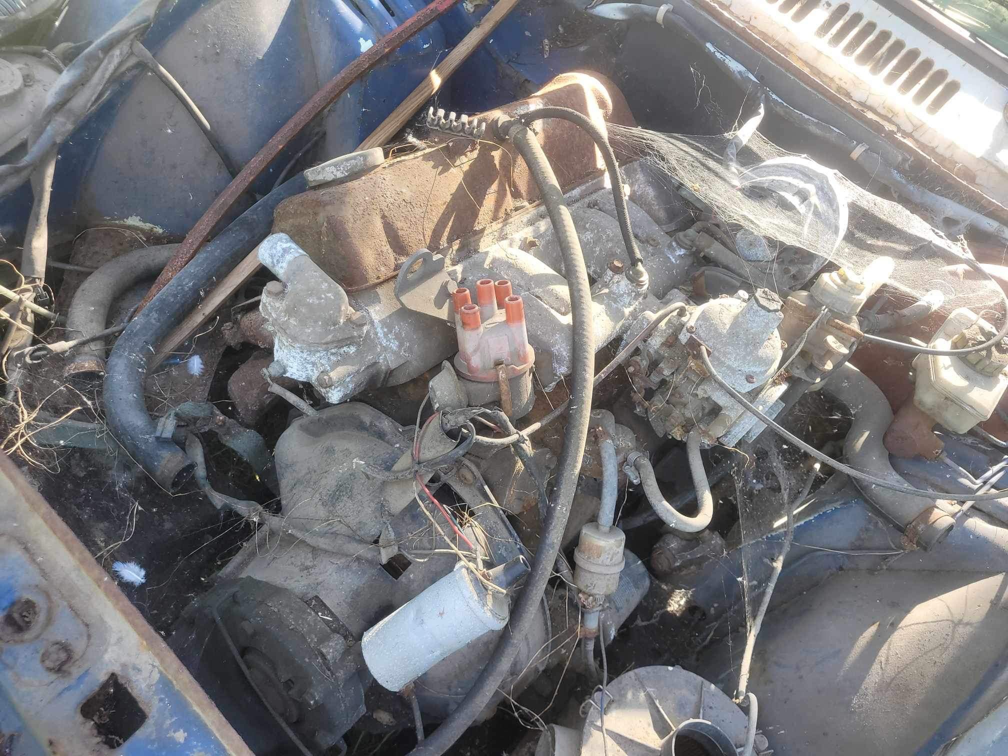 Zardzewiały i zabrudzony silnik samochodowy, częściowo zdemontowany, widoczny pod otwartą maską, pokryty pajęczynami i kurzem.